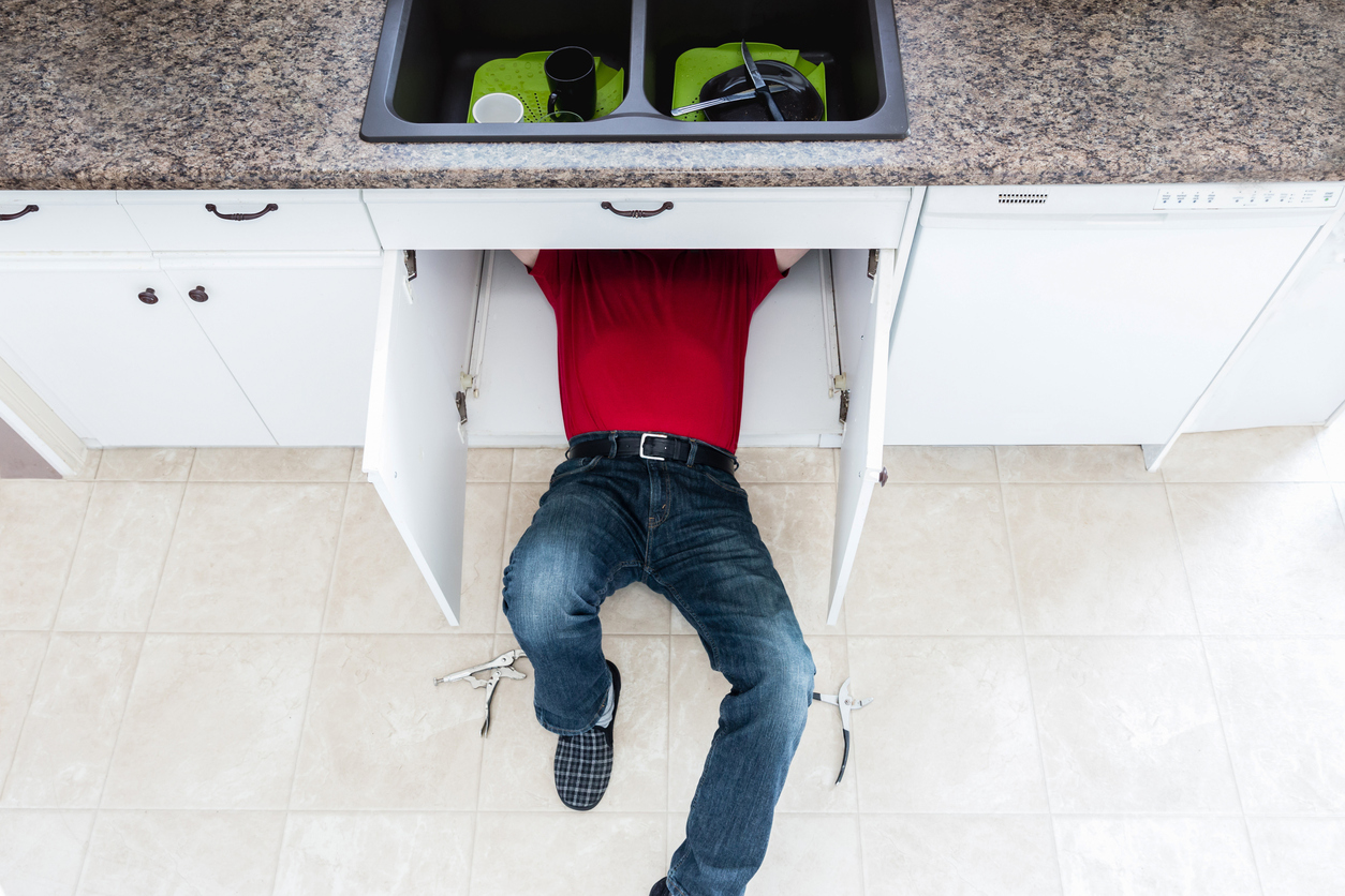 horizontal image of a man in a red top and jeans lying under the kitchen sink cabinet and fixing a leak